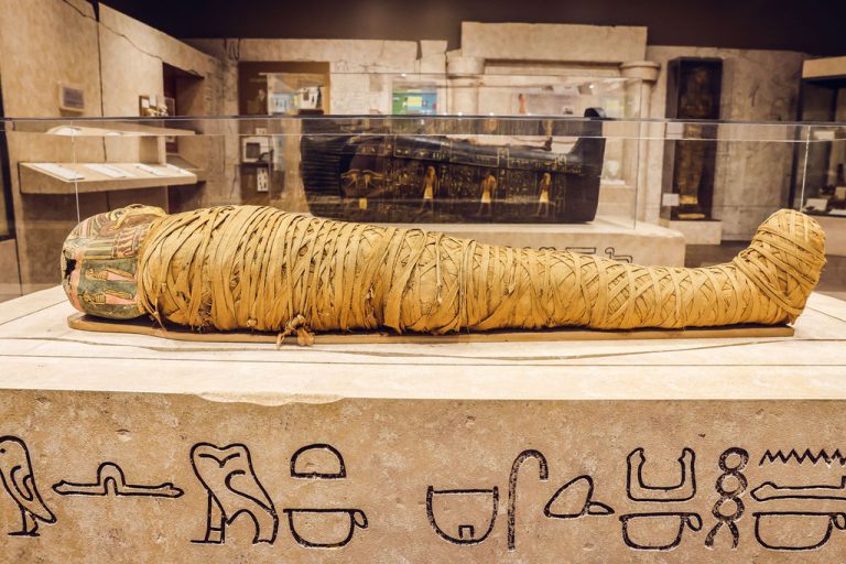 Unearthing Ancient Egypt gallery as if you were about to walk in. Pictured at the forefront is a mummy wrapped in traditional linens dating back to 332 BC. In the background is a black coffin with intricate design and art that dates back to between 1479 and 1336 BC.