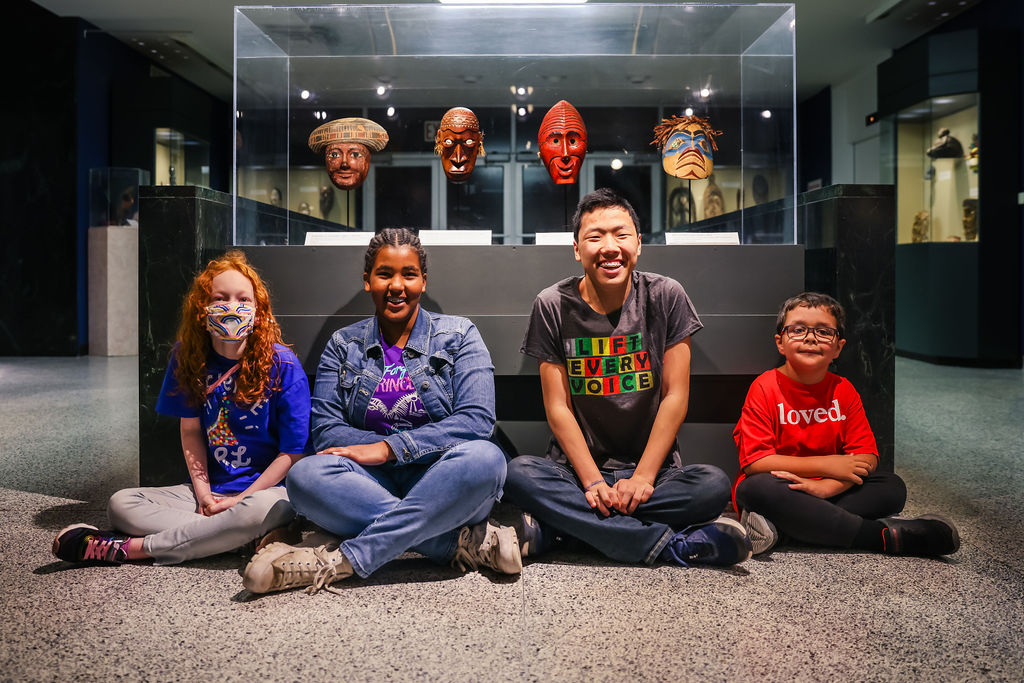 Four kids are sitting in front of four masks which are part of the Face From Our Past exhibit.