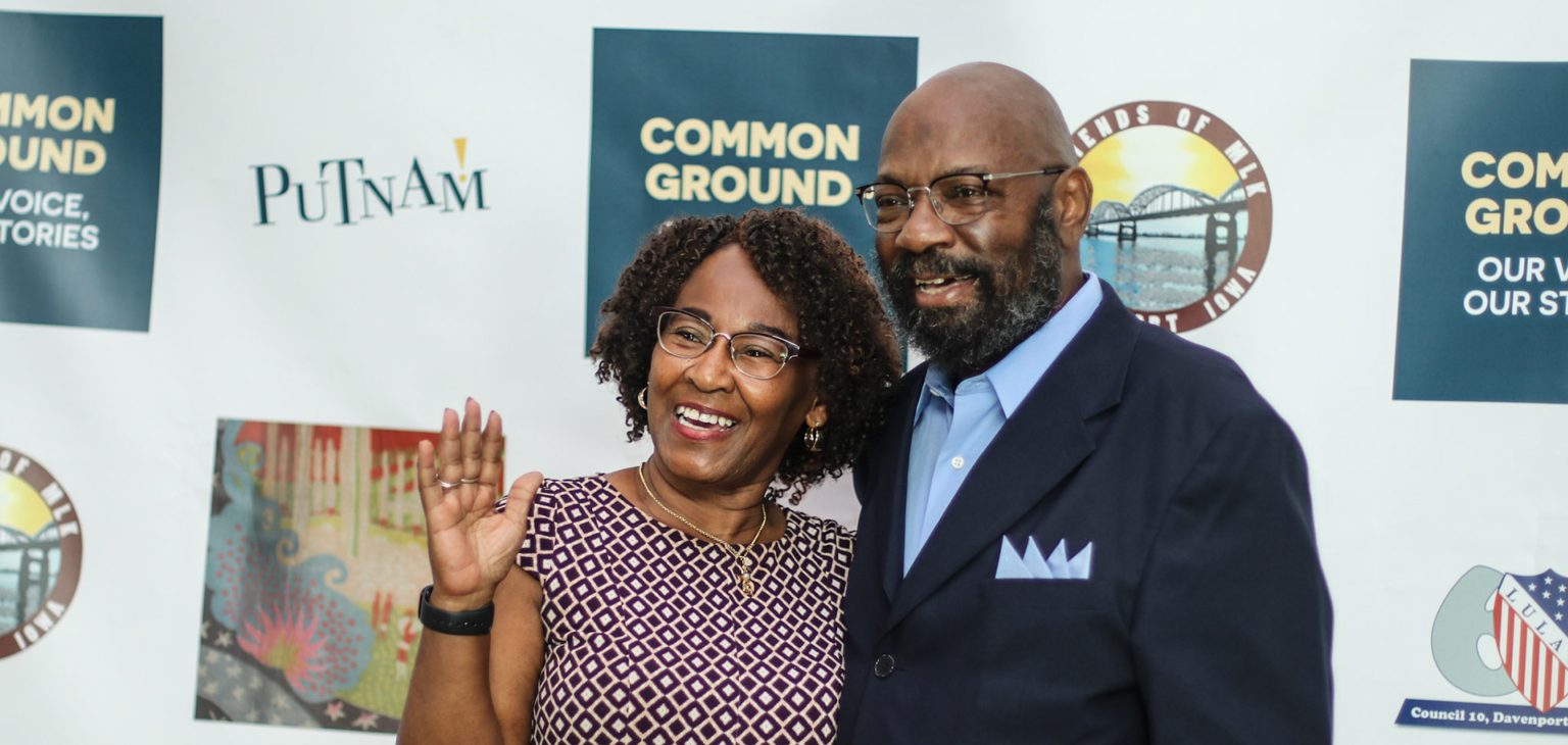 Business Partnerships. Featuring African American woman with glasses and patterned dress and African American man with glasses and in blue suit with a light blue shirt attending an event in front of a Common Ground, Putnam photo background.