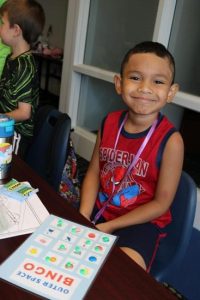A young boy sitting at a desk wearing a spider man shirt looks up from his paper and smiles at the camera.