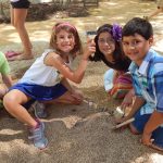 Muti-cultural children in brightly colored shirts digging for replica dinosaur fossils, with brushes in the outdoor Dino Dig area at the Putnam Museum.