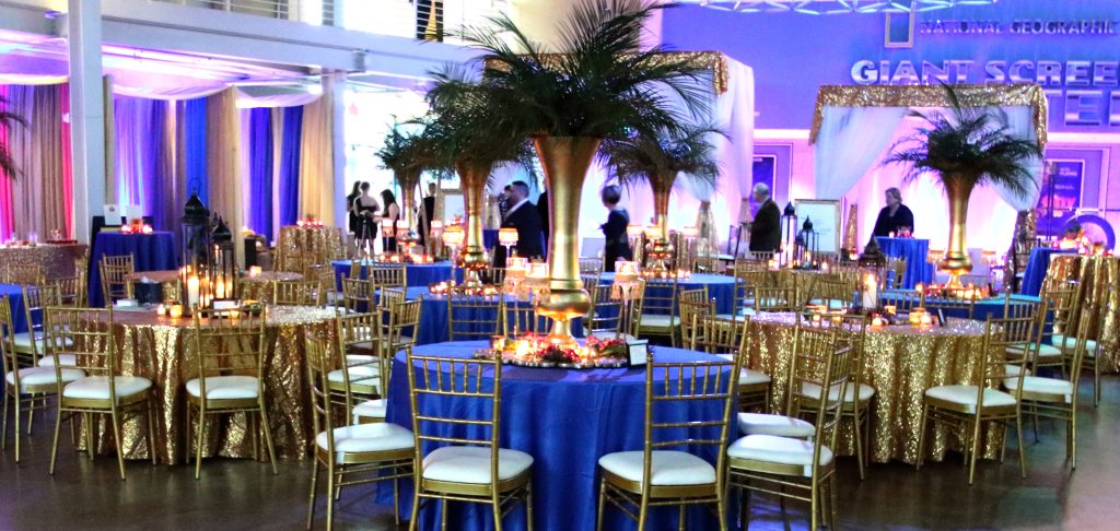 Grand Lobby at the Putnam Museum decorated with gold chairs, tall golden vases with palms, purple tablecloths and dramatic purple and pink lighting.