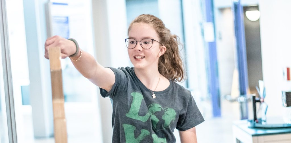 Pictured here is a young woman, approximately 14 years old, smiling as she pulls a lever on an interactive in the Putnam’s Science Center. She is wearing glasses and a t-shirt that spells the word “LOVE” with an Earth representing the letter ‘O.’