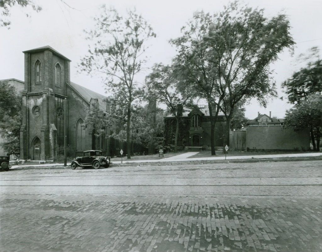 Black and white photo of the original location of the Putnam Museum, featuring tower on a cobblestone street with a historic car parked on the street.
